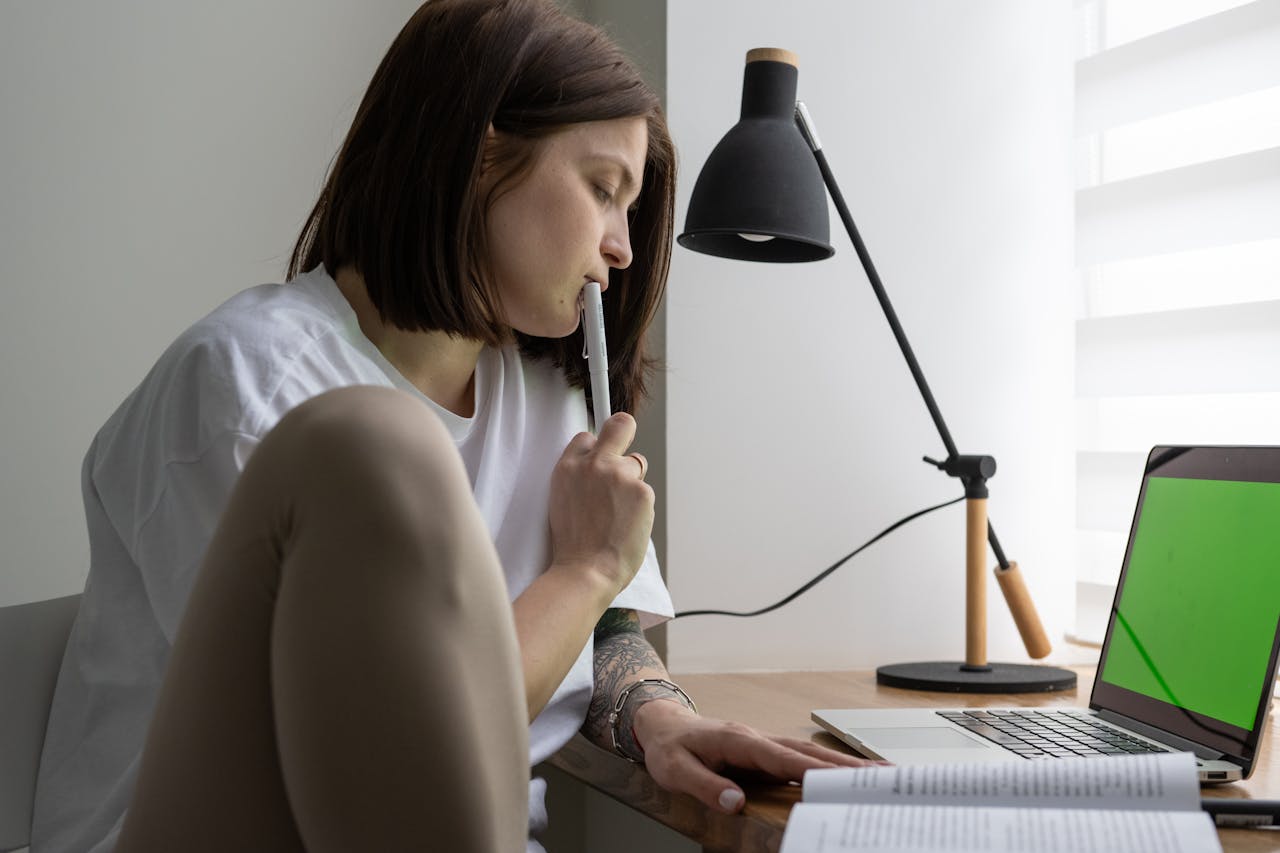 Woman studying at home using a laptop and book, deep in thought during the day.